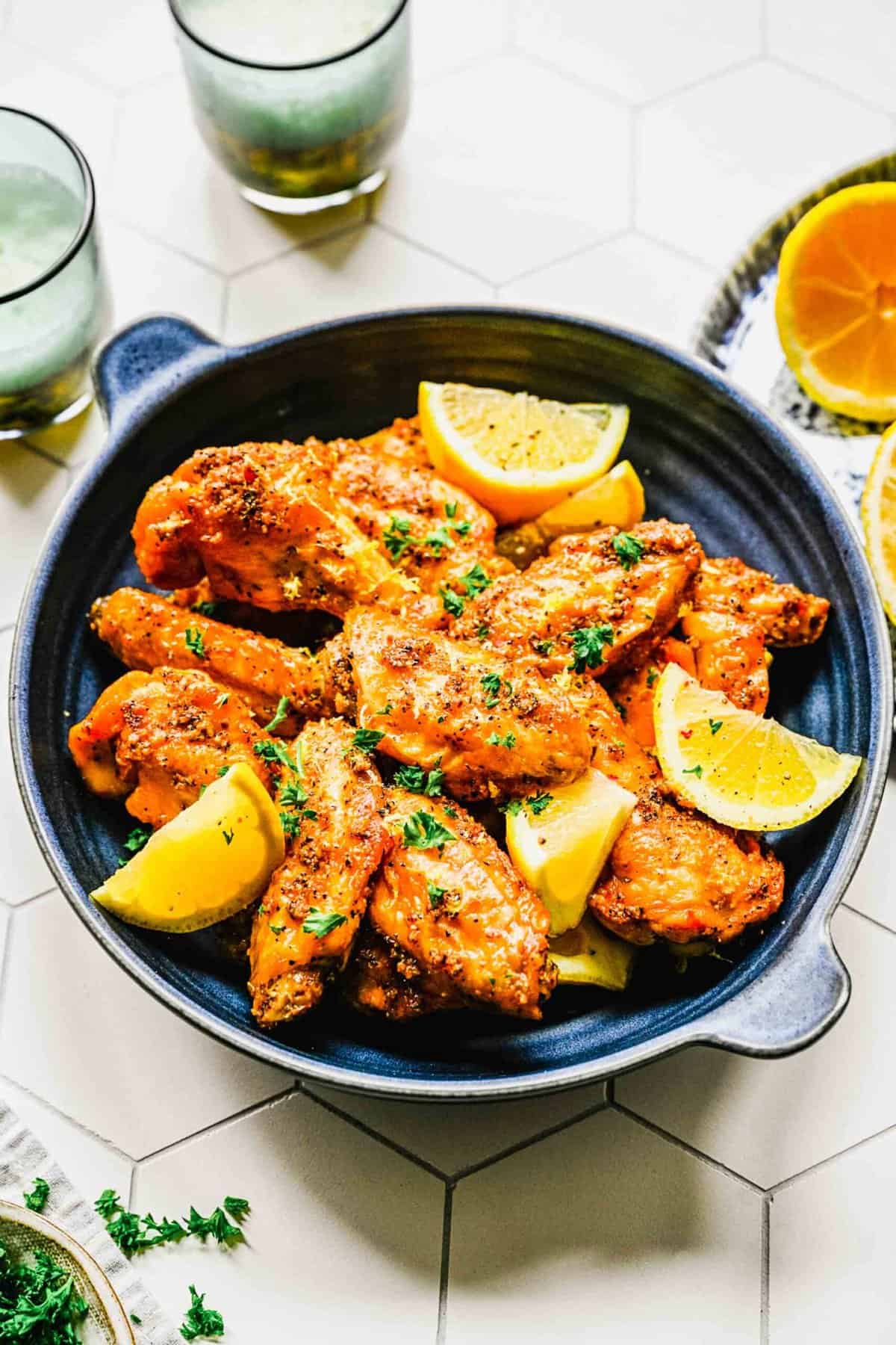 Lemon pepper chicken wings on a platter with lemon wedges near 2 drinking glasses.