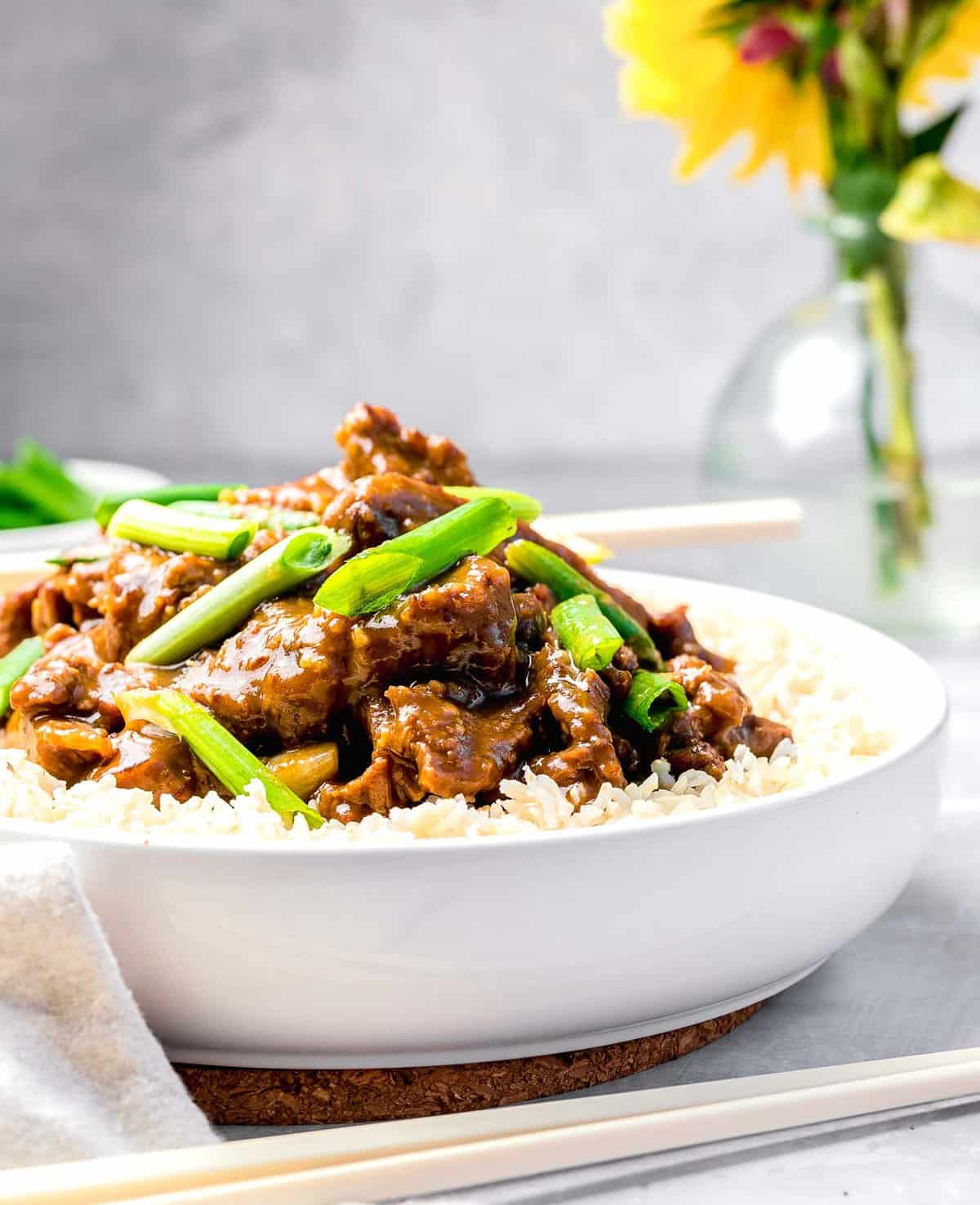 Mongolian beef served in a bowl over rice next to flowers and chopsticks.
