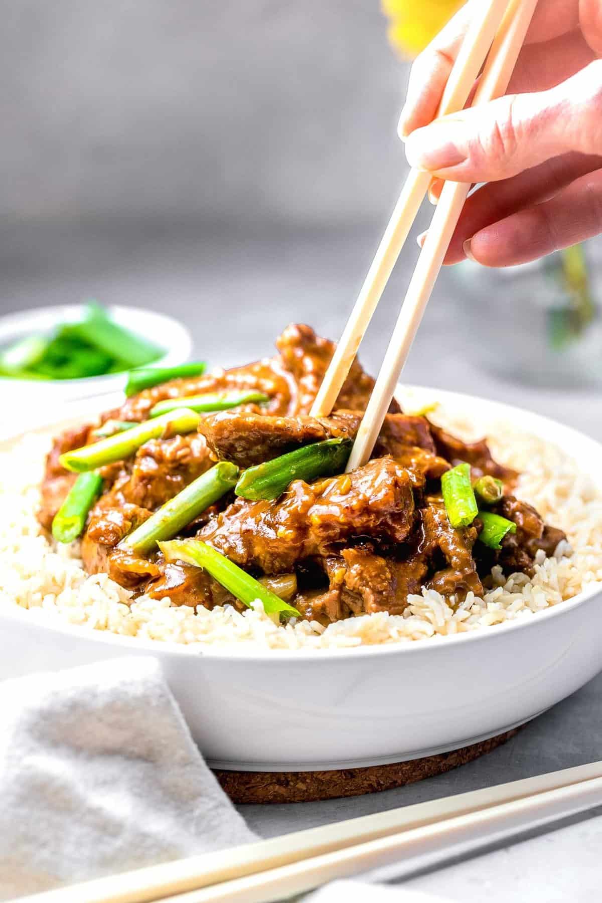 Mongolian beef served in a bowl over rice with a hand wielding chopsticks.