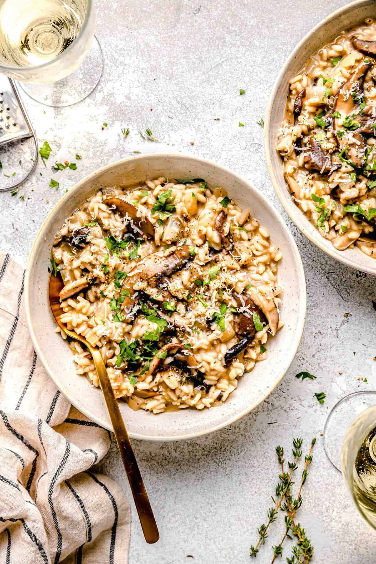 Mushroom risotto in dinner bowls with a spoon next to a glass of white wine and garnished with fresh parsley.