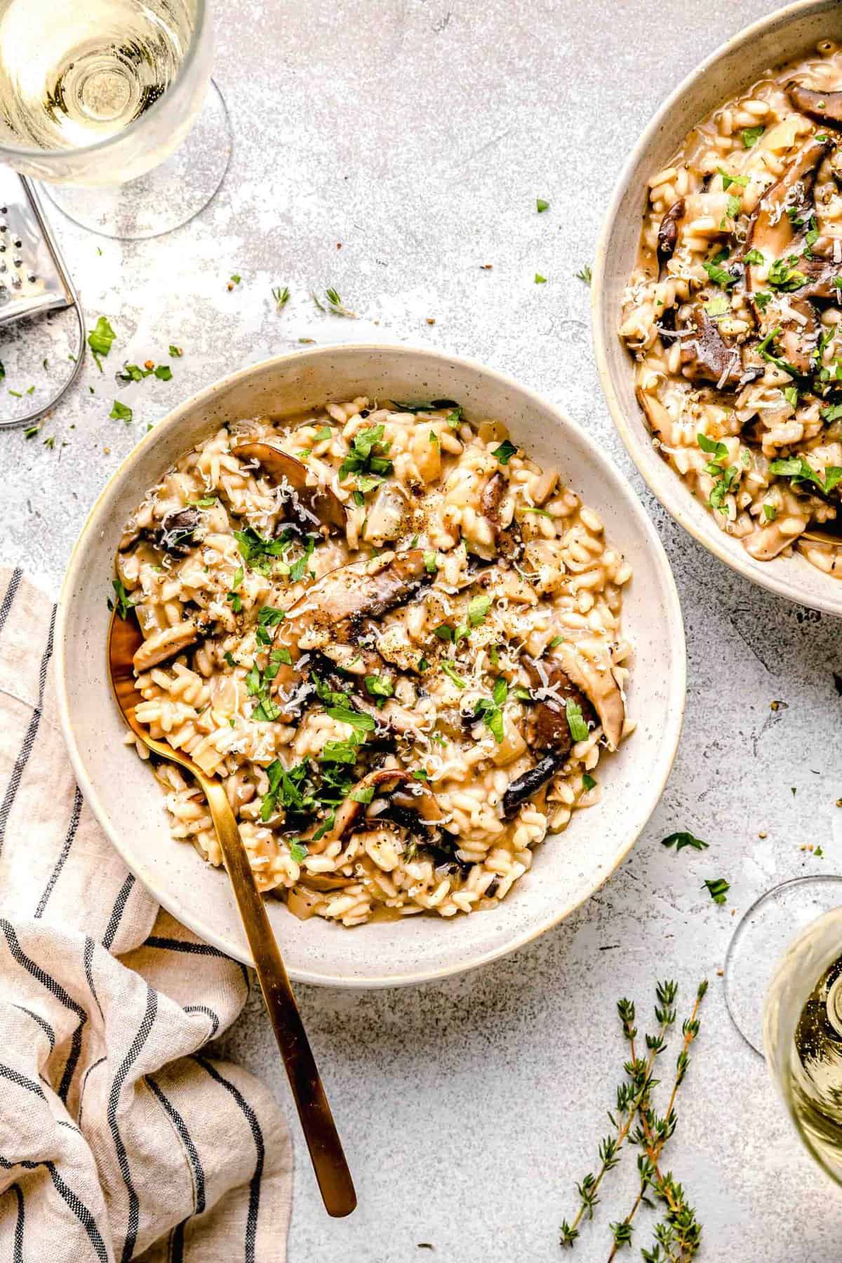 This mushroom risotto marries two kinds of mushrooms, arborio rice, and nutty parmesan cheese for a deeply savory, delightfully creamy dish. Mushroom risotto in dinner bowls with a spoon next to a glass of white wine and garnished with fresh parsley.