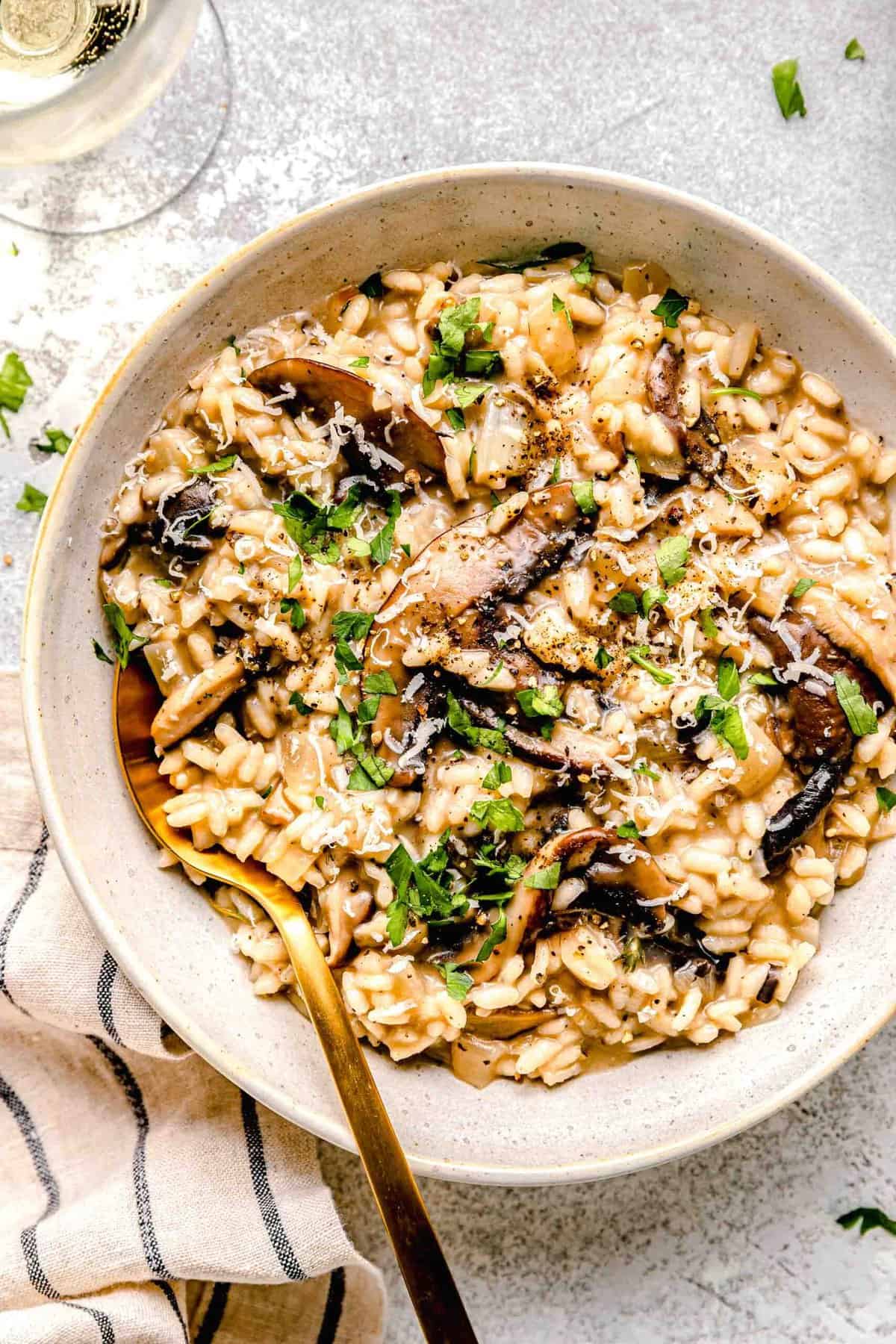 Mushroom risotto in a dinner bowl with a spoon next to a glass of white wine and garnished with fresh parsley.