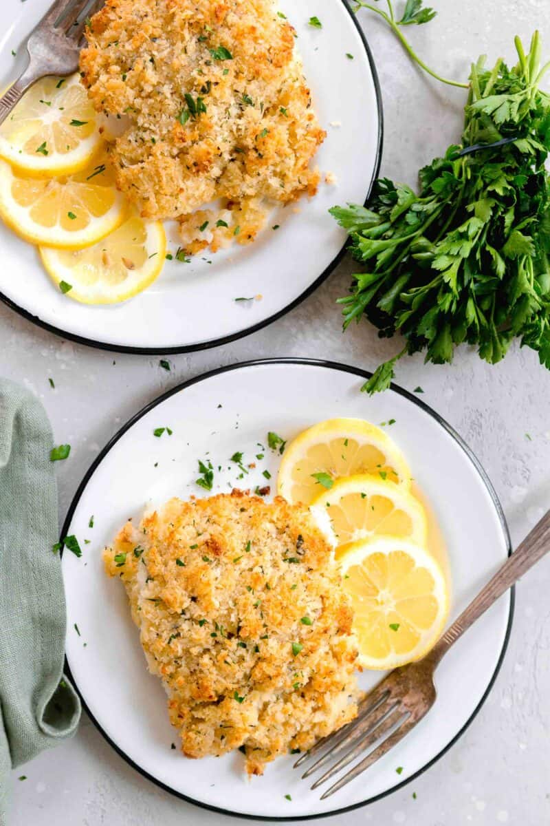 two black rimmed white plates with cooked panko parmesan cod and lemon slices on the plate with antique forks. fresh parsley bunch and green linen towel can be seen next to the plates