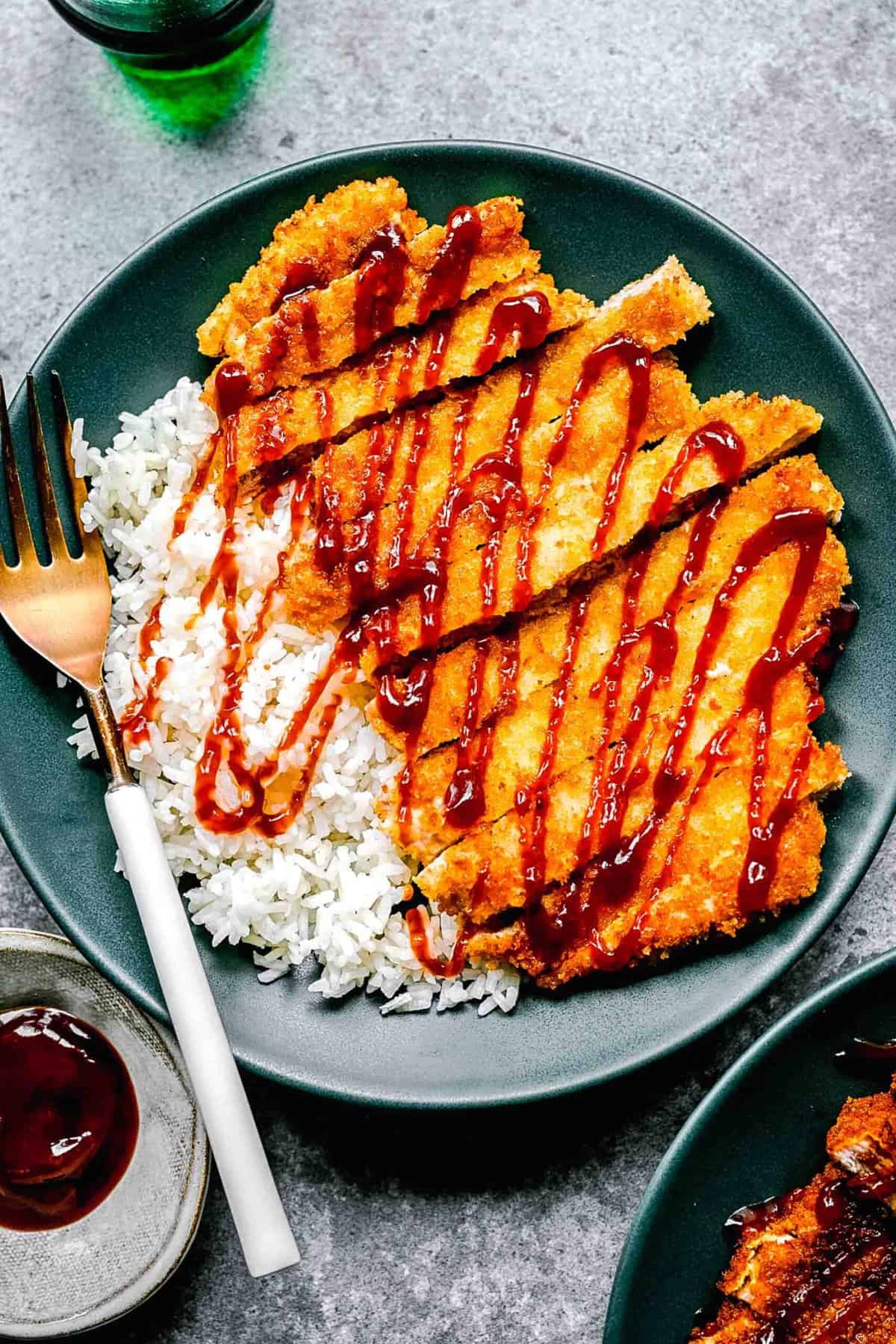 Pork katsu on a plate with a fork over white rice and sauce.