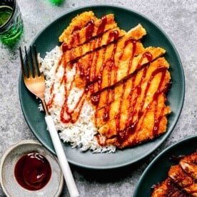 Pork katsu on a plate with a fork over white rice and sauce next to sauce and drinking glasses.