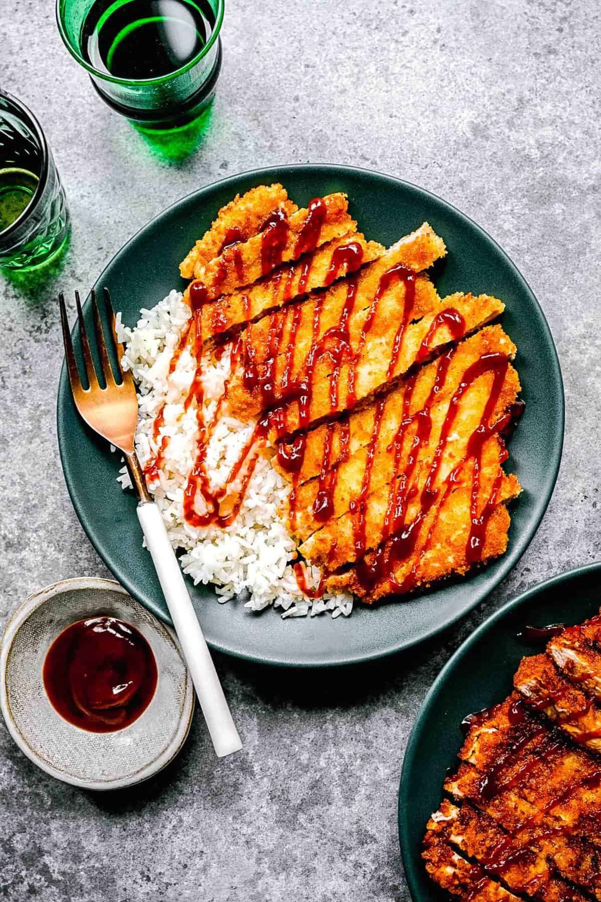 Pork katsu on a plate with a fork over white rice and sauce next to sauce and drinking glasses.
