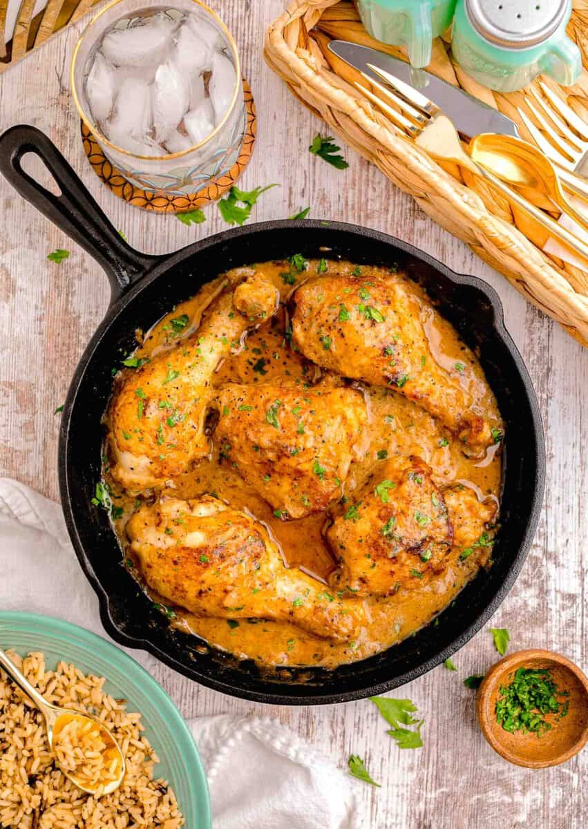 overhead image of smothered chicken in a cast iron skillet next to a glass of water and tray of utensils and plate of rice