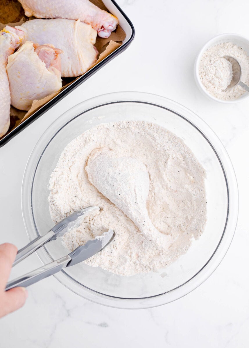 coating chicken leg in a large bowl of flour mixture next to a rimmed baking sheet of raw chicken