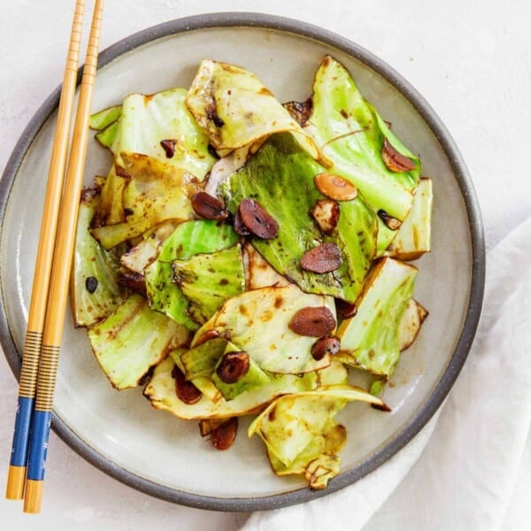 stir fry cabbage on a ceramic grey plate with soy sauce coating the cabbage leaves and garlic slices. wooden chopsticks can be seen on the plate and next to a linen towel