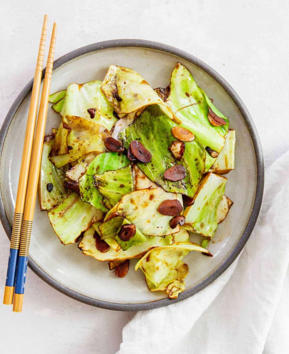 stir fry cabbage on a ceramic grey plate with soy sauce coating the cabbage leaves and garlic slices. wooden chopsticks can be seen on the plate and next to a linen towel