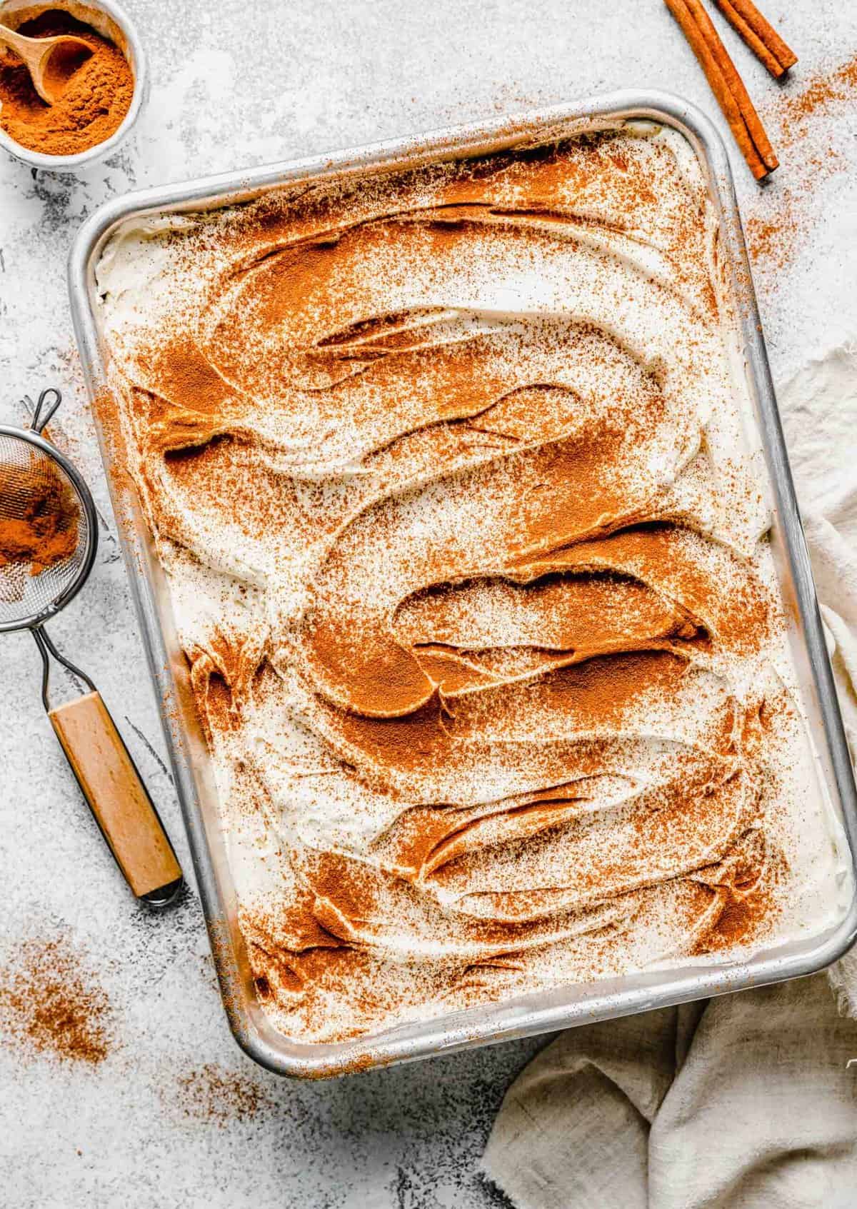 Prepared tres leches cake near a bowl of cinnamon, cinnamon sticks, and a sifter.