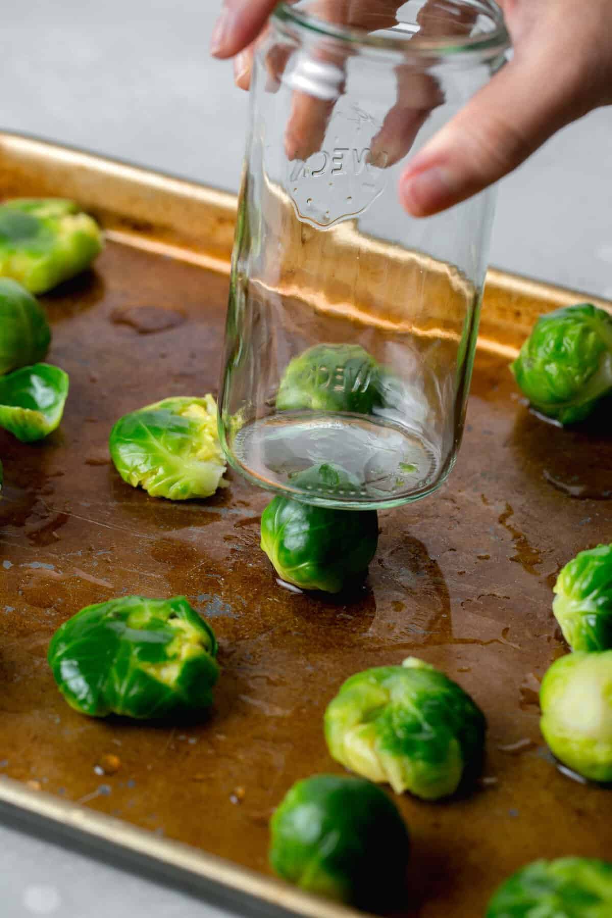brussels sprouts on a rimmed baking sheet with a flat bottom glass being pushed on top of a brussel sprout