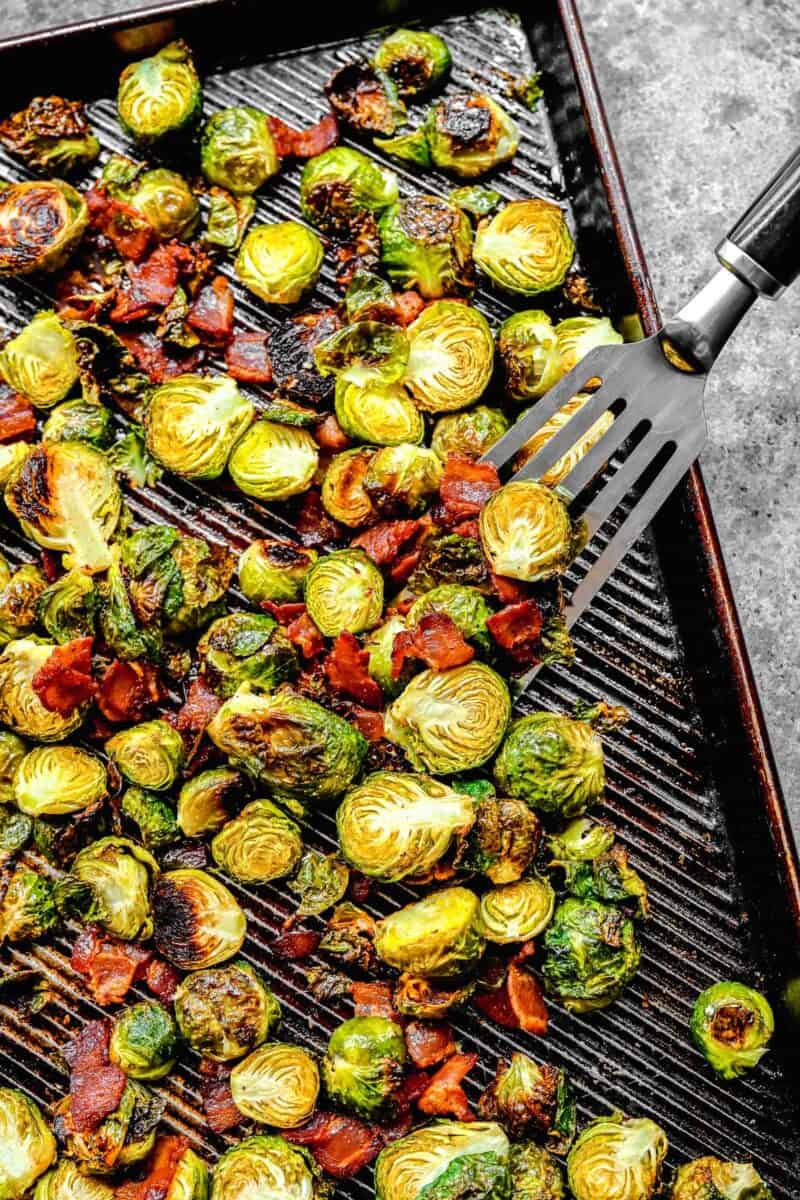 a metal fish spatula scooping bacon and brussels sprouts on a rimmed baking sheet