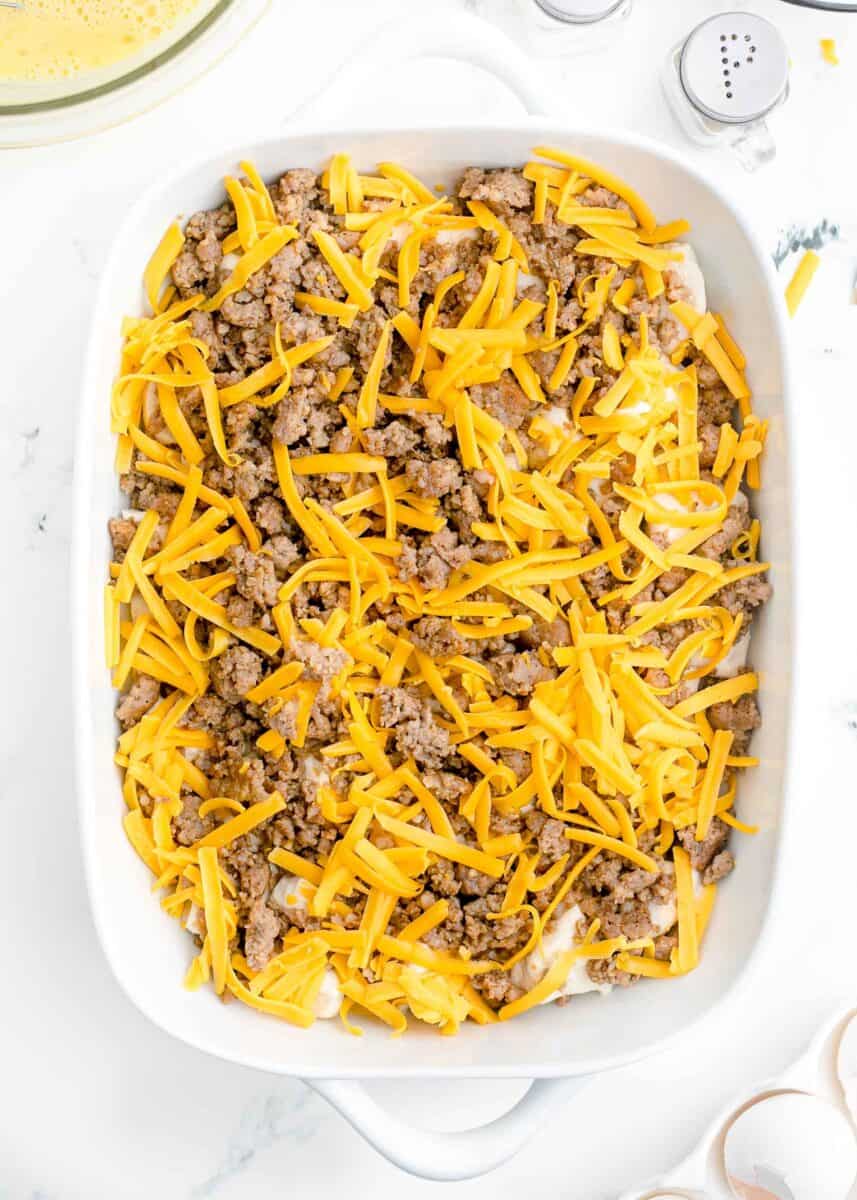 shredded cheese sprinkled on top of browned sausage and biscuits in a white baking dish