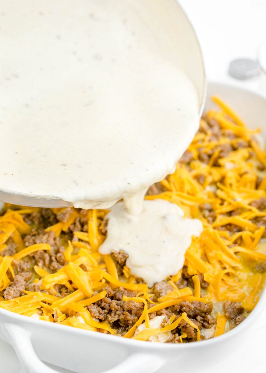 thickened white gravy being poured on top of baking dish with browned sausage and shredded cheese