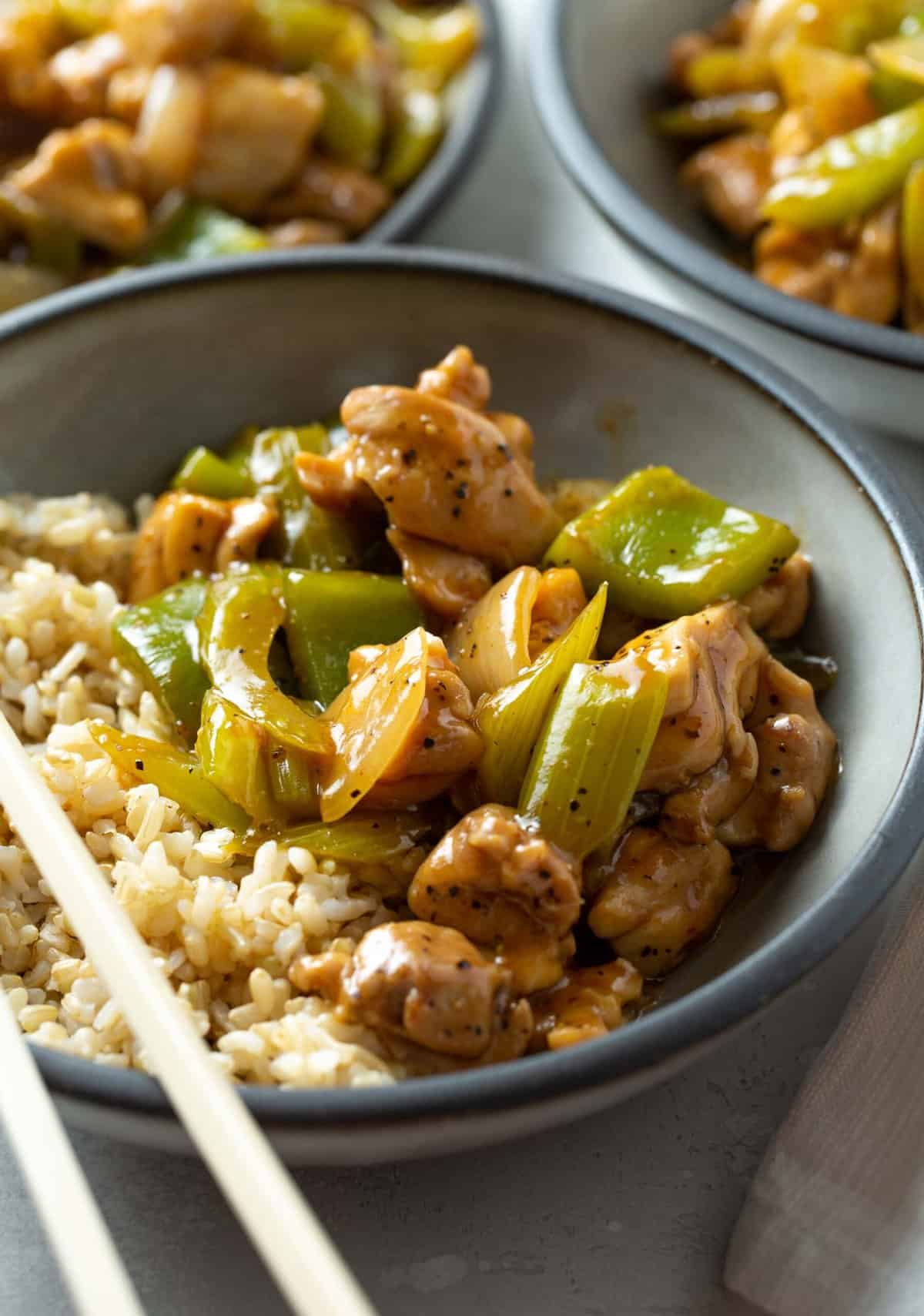 black pepper chicken in a grey bowl with brown rice with chopsticks laying on the bowl