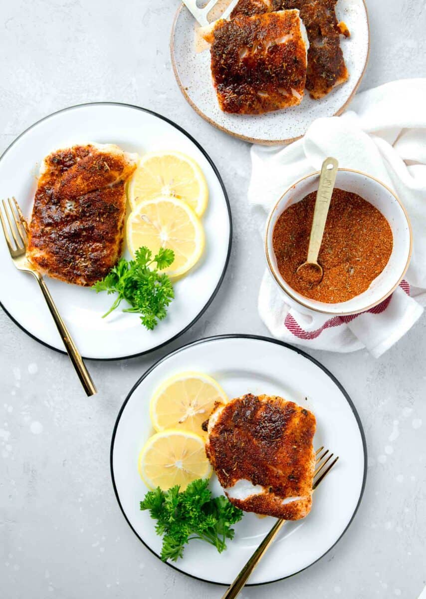 blackened cod on white and black rimmed plates with lemon slices and parsley next to a bowl of blackening seasoning