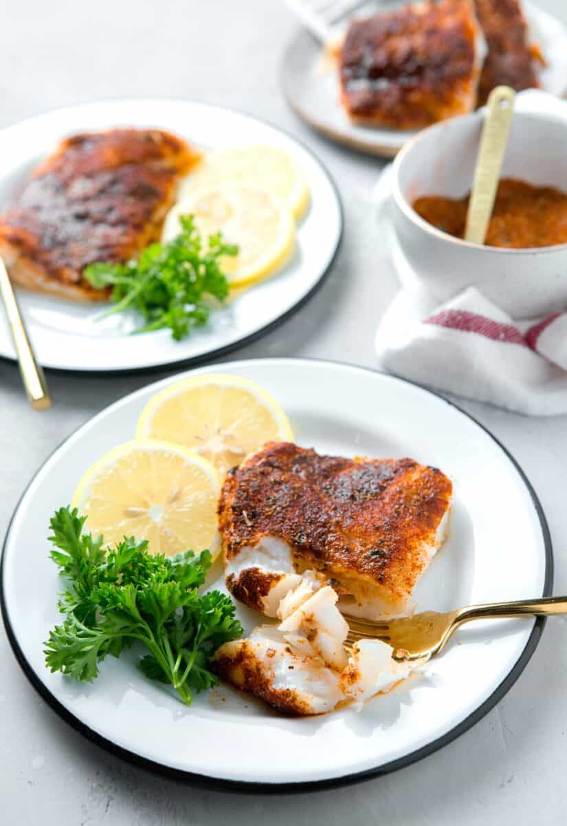 cod filet on a white and black rimmed plate with a gold fork piercing the flesh next to lemons and parsley