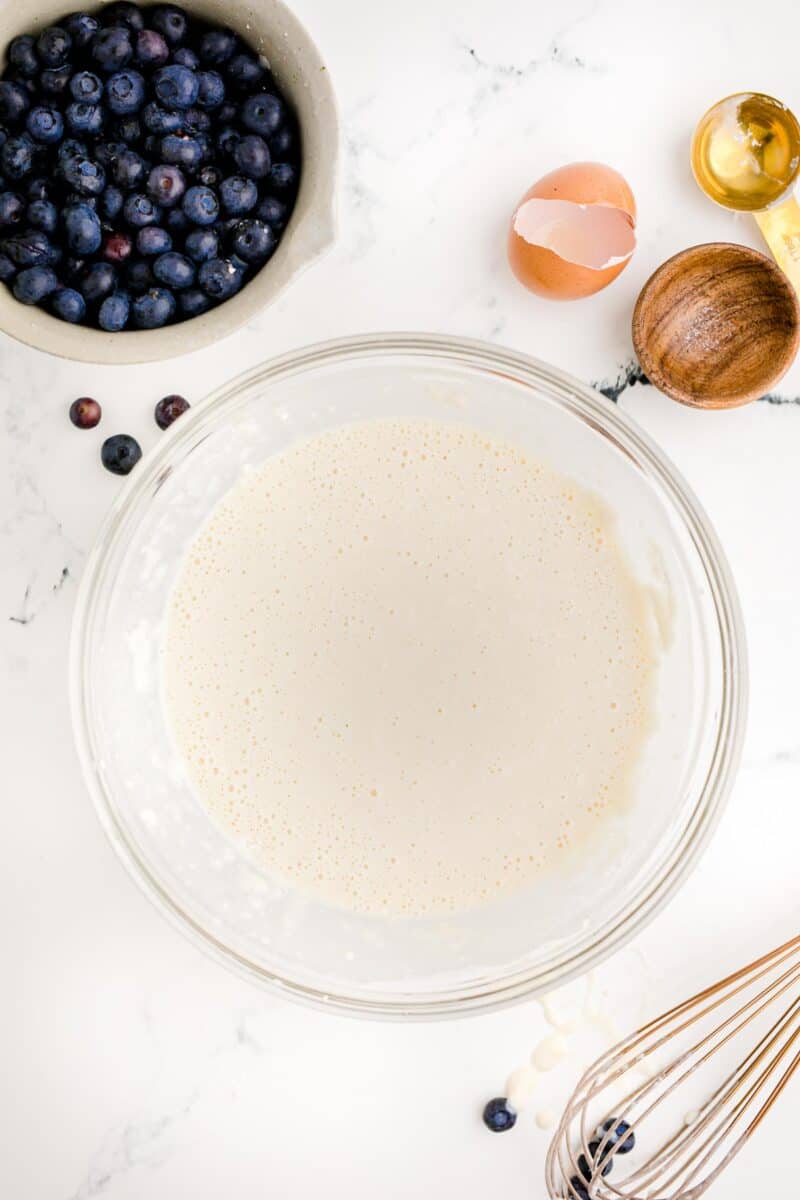 a bowl of pancake batter is presented on a white surface next to a bowl of blueberries.