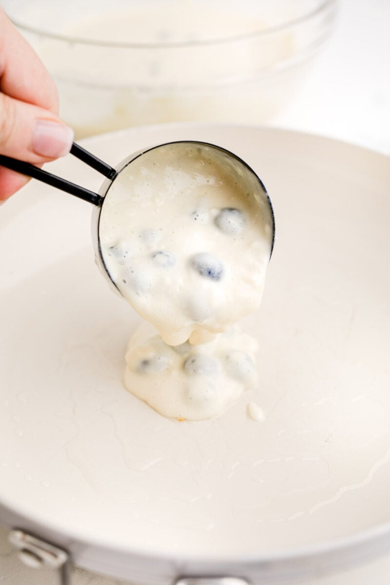 blueberry pancake batter is being poured into a white skillet.
