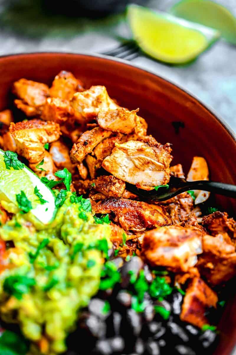 chicken, beans, and guacamole are placed on top of white rice in a brown bowl.