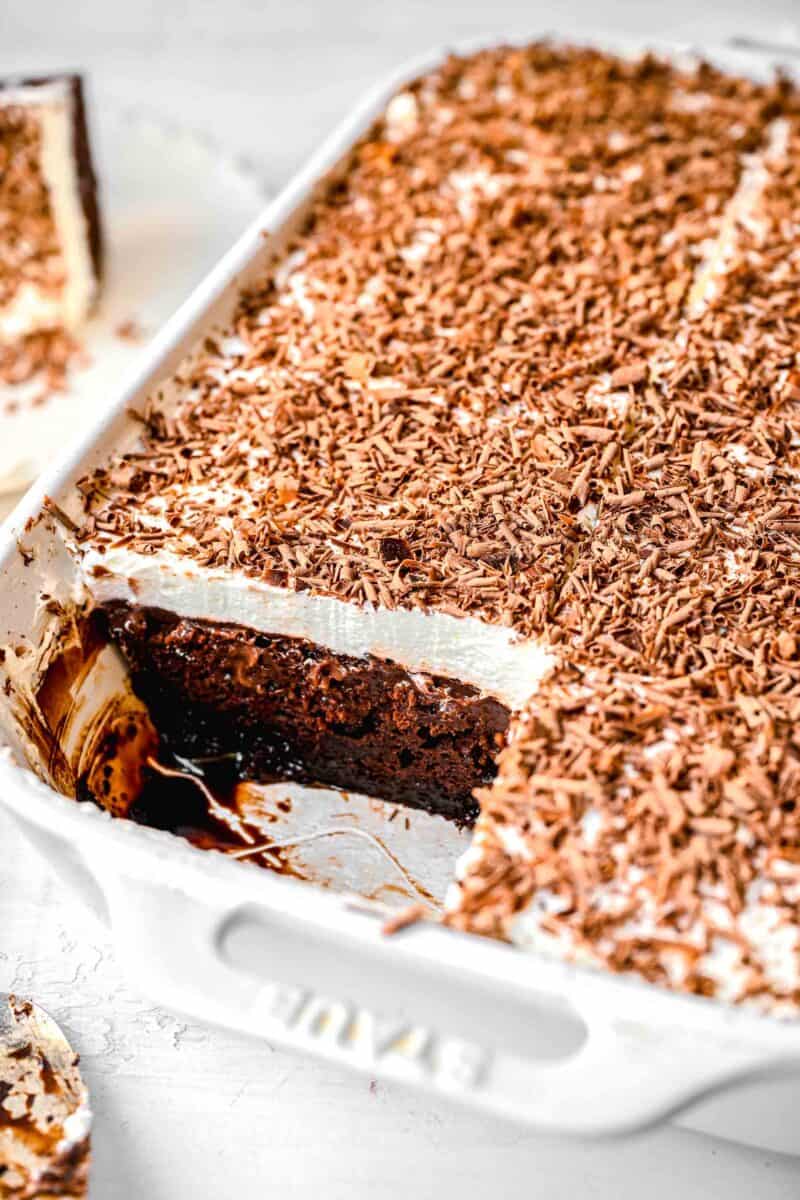 a slice of chocolate pudding cake taken out of the baking dish to show the inside of the cake. there is whipped topping with a lot of chocolate shavings on top of the cake in the white baking dish