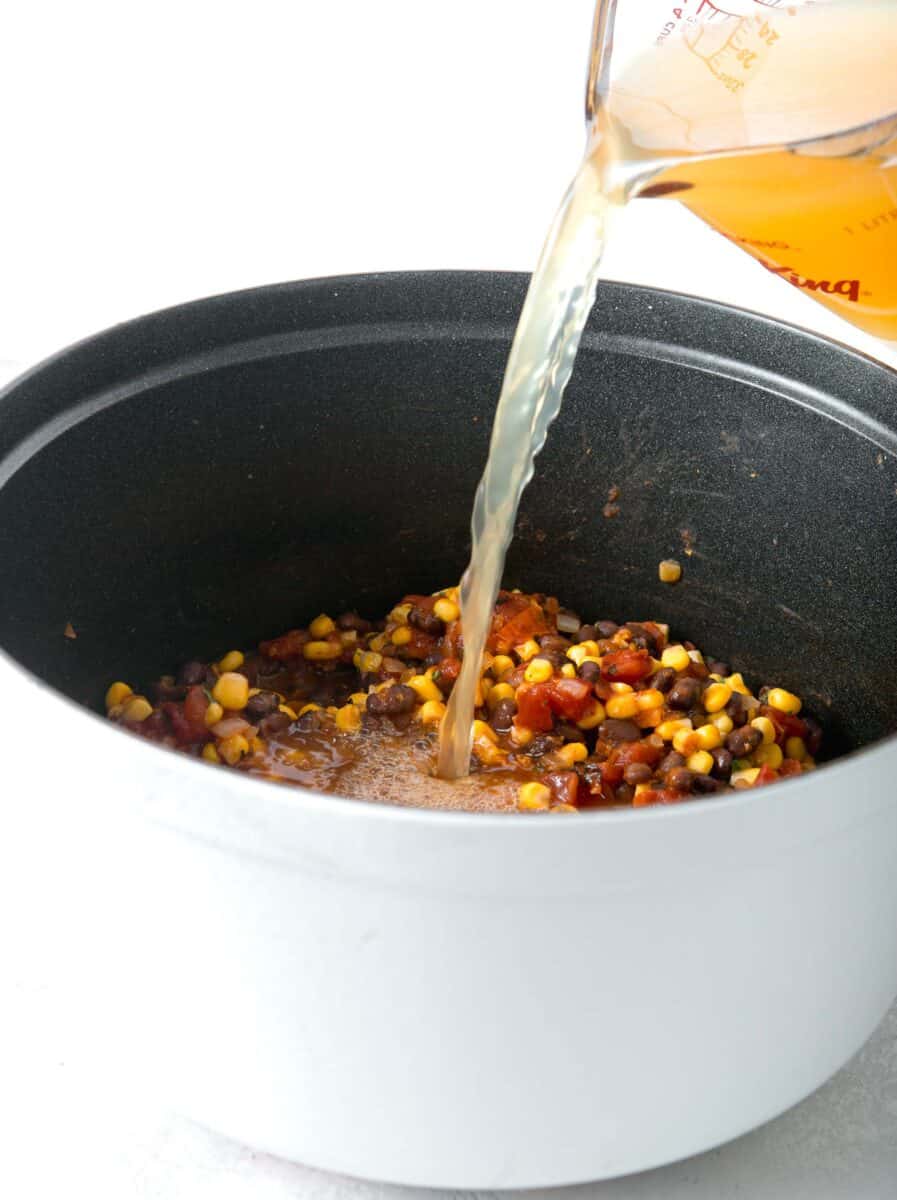 chicken broth being poured into the pot with black beans, diced tomatoes, and corn