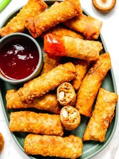 Crispy golden spring rolls on a plate with dipping sauce; one spring roll cut in half and another dipped in sauce.