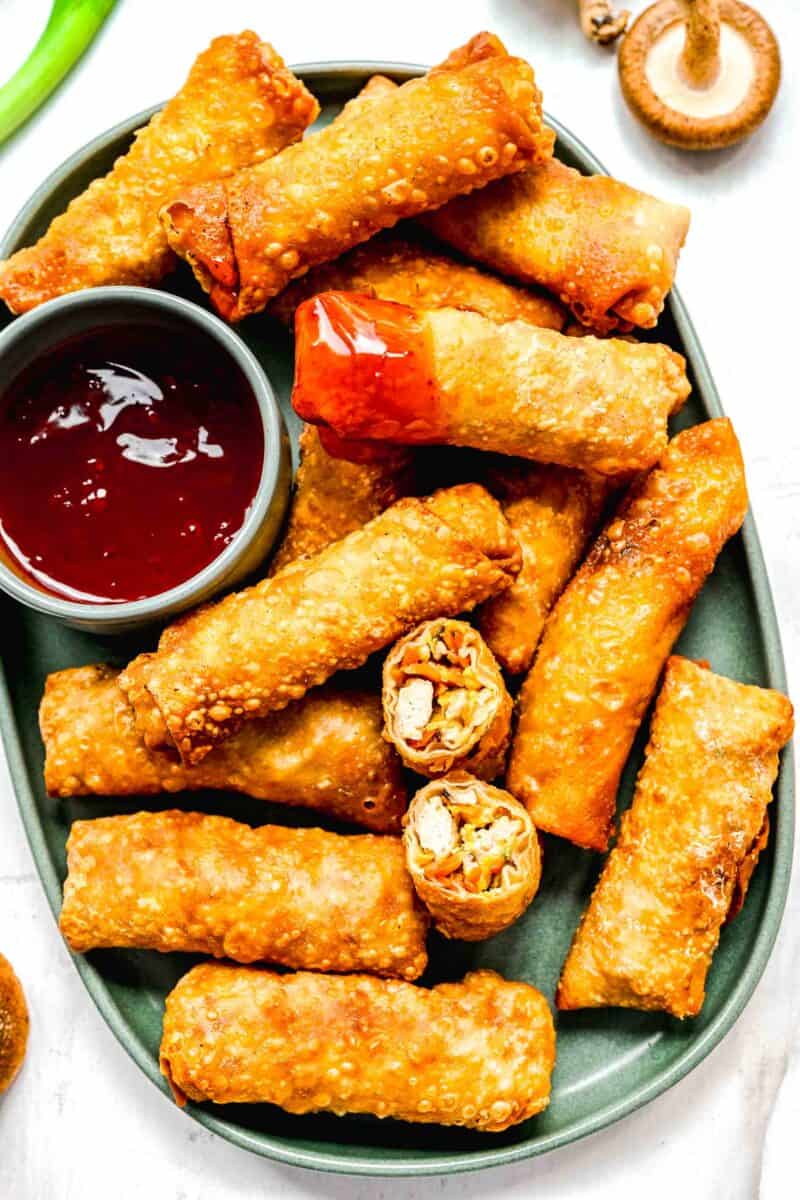 Crispy golden spring rolls on a plate with dipping sauce; one spring roll cut in half and another dipped in sauce.
