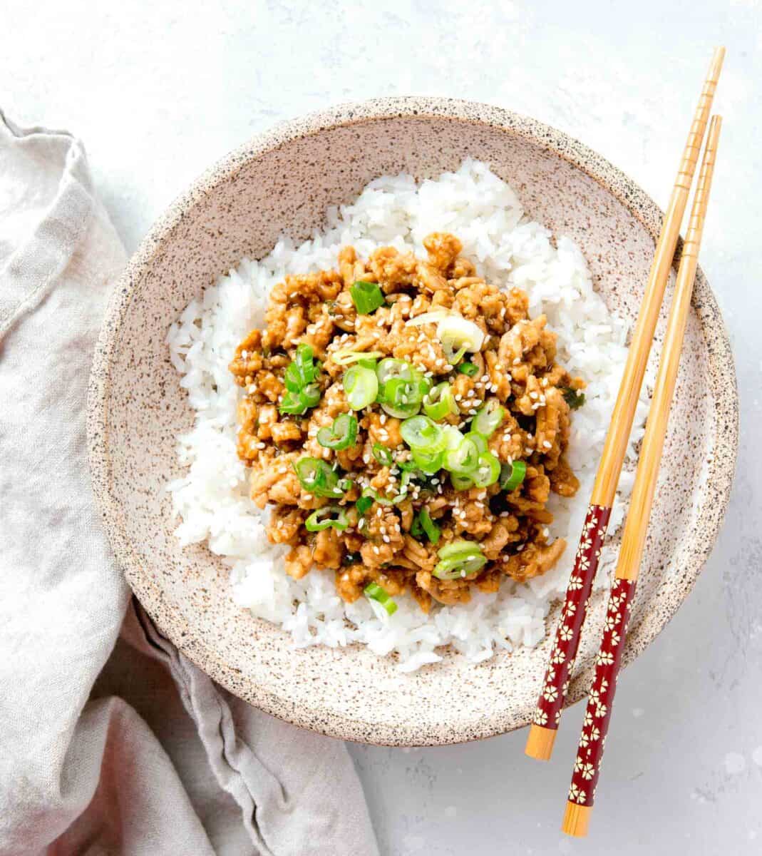 ground chicken teriyaki in a speckled brown bowl on top of white rice. wooden chopsticks are laying on top of the bowl which is next to a linen towel