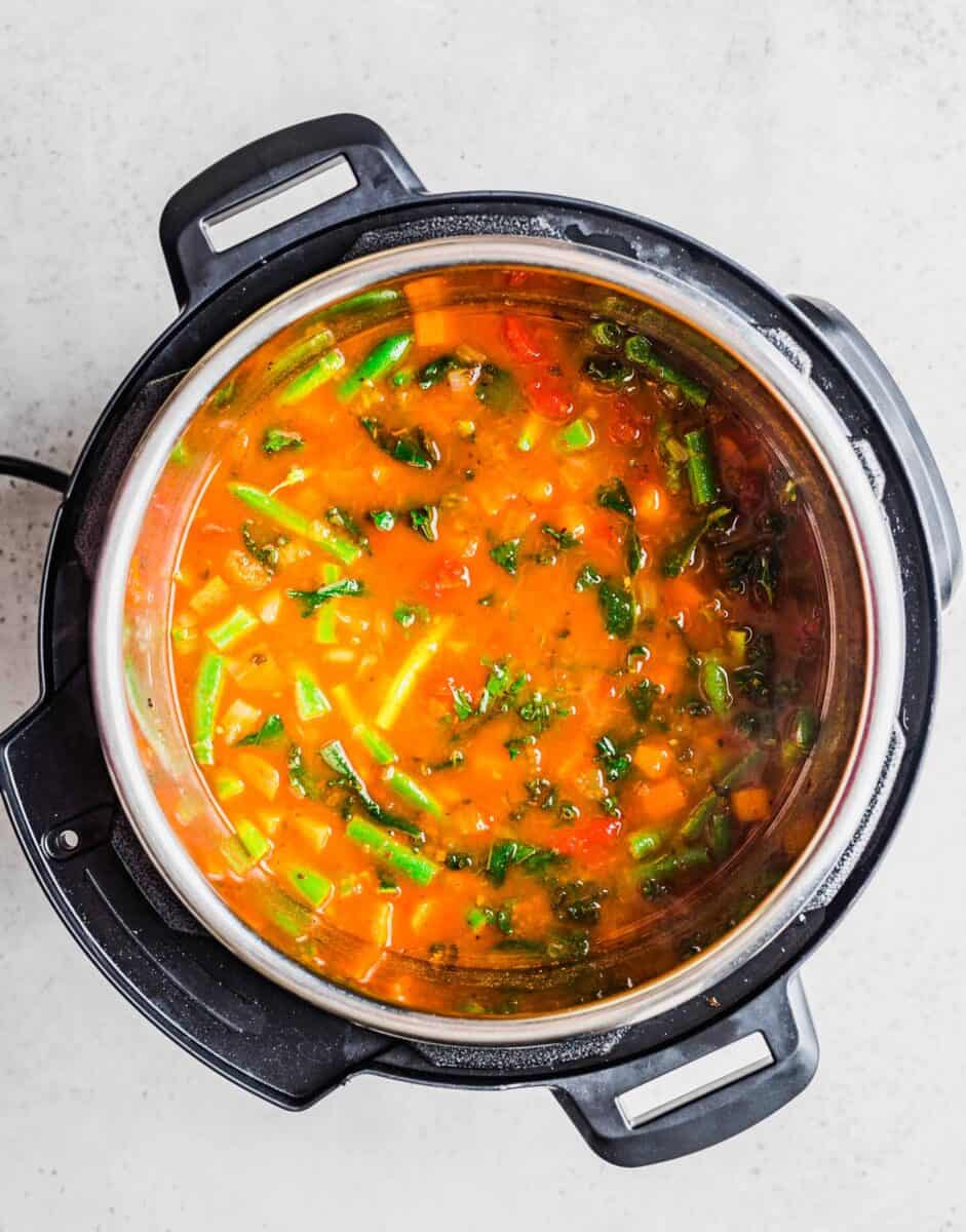 vegetables are stirred into a red broth filling an instant pot.