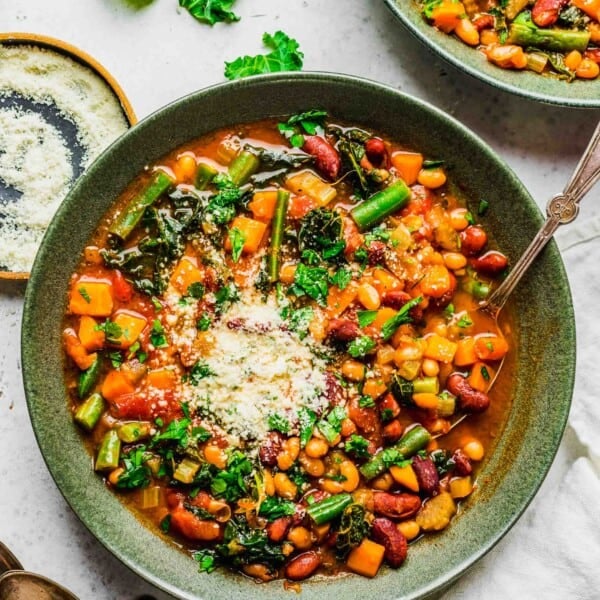 an overhead image shows the vibrant colors of the beans and vegetables in the soup. a spoon can be seen in the green bowl with the soup along with fresh grated parmesan on top