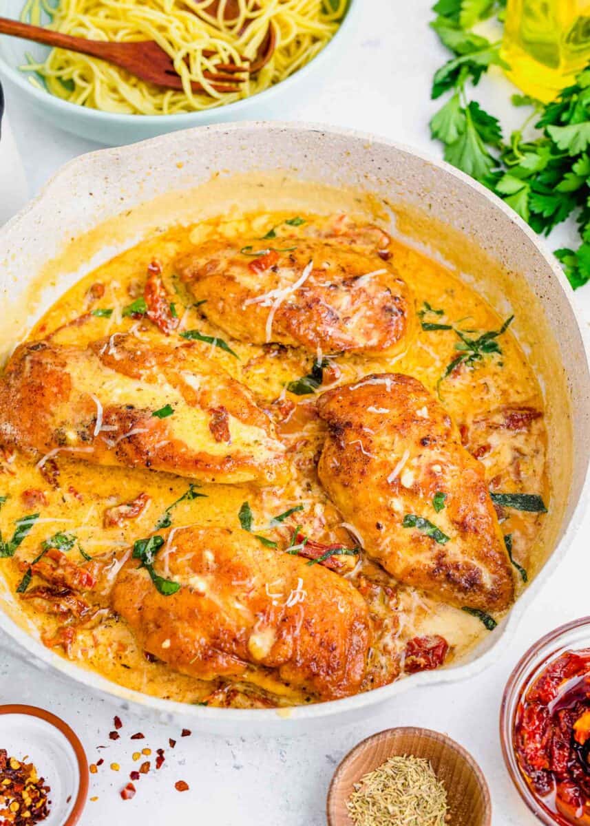 seared chicken breast in cream sauce mixture in a skillet with parmesan cheese and basil on top. a bowl of pasta can be seen in the background