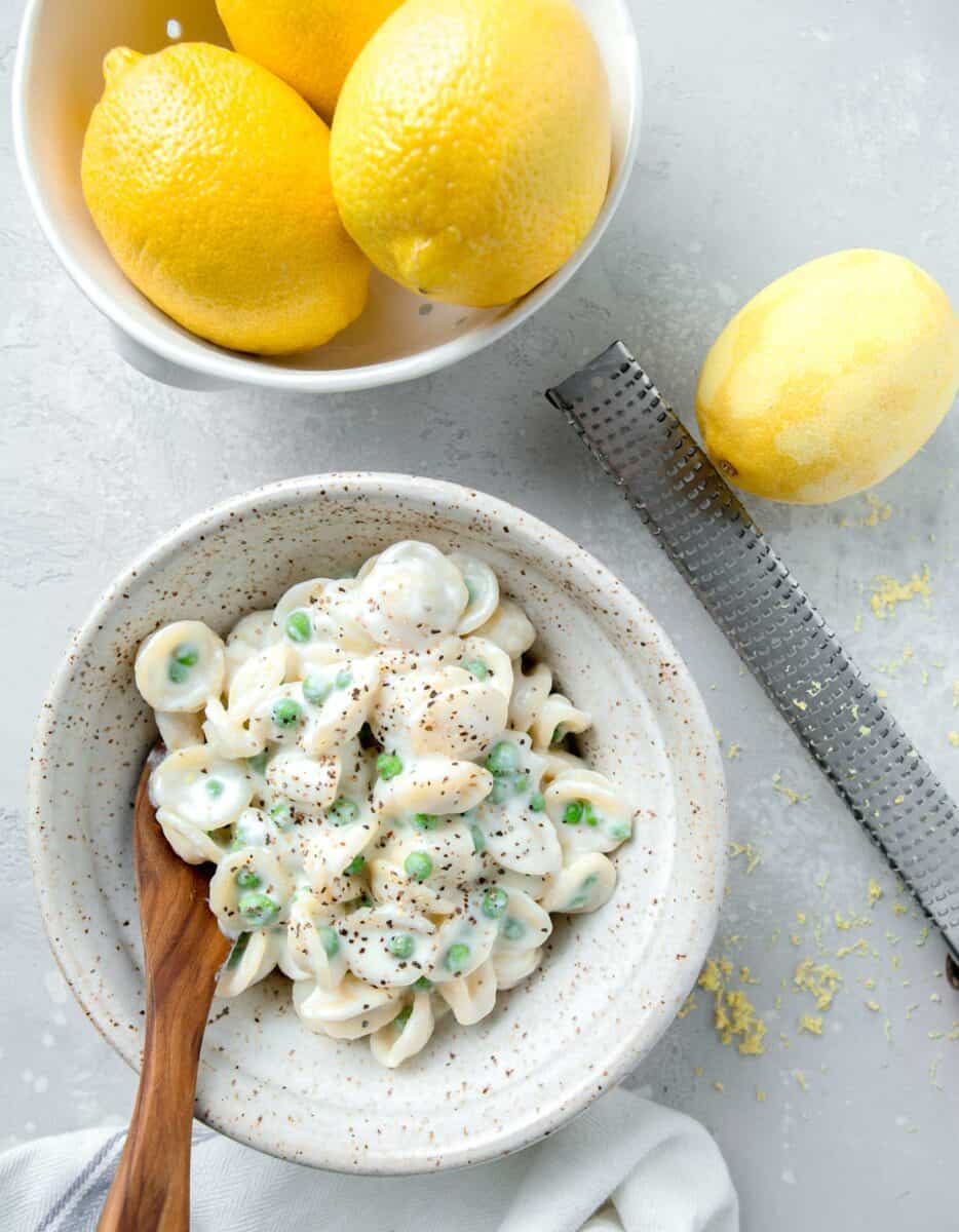 overhead image of ricotta and lemon pasta with peas. the pasta is in a speckled bowl with a wooden spoon next to a lemon zester and lemons in a colander.