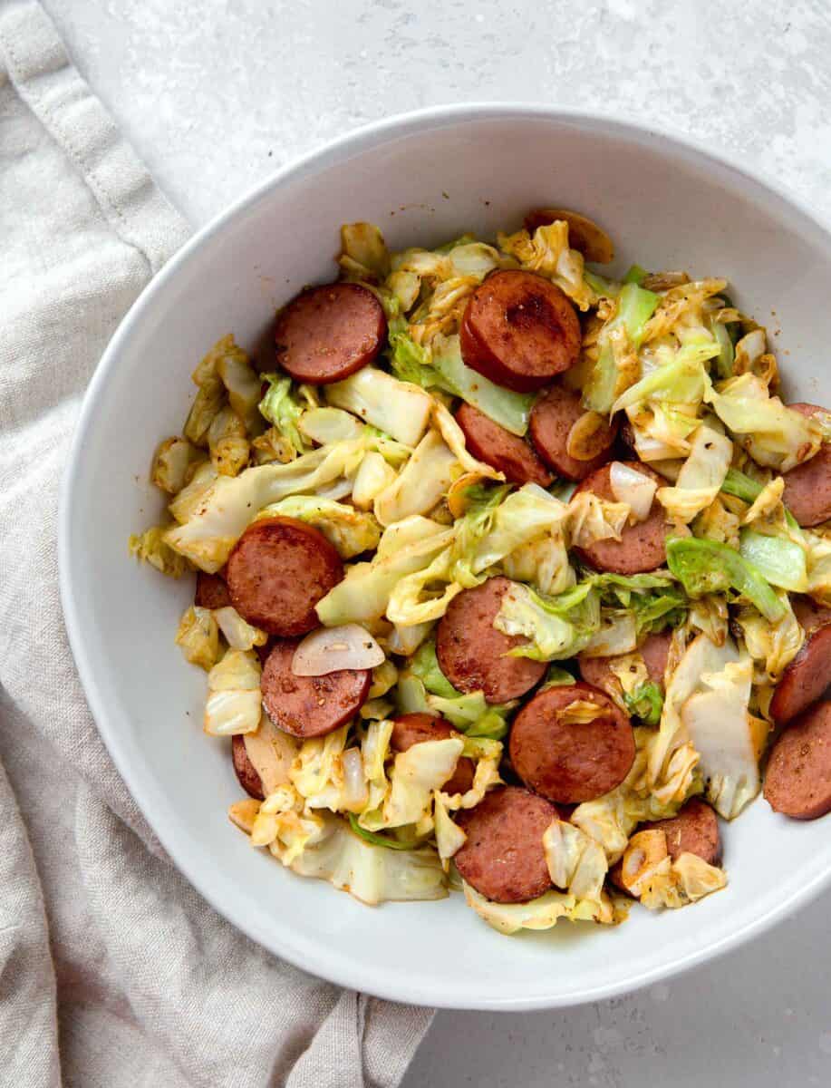 sauteed cabbage and sausage in a white bowl next to a linen towel