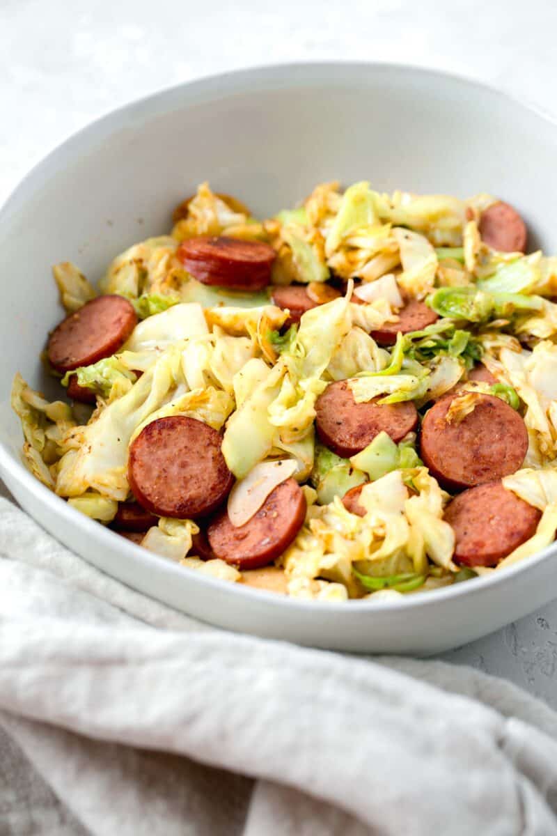 sauteed cabbage and sausage in a white bowl next to a crumpled linen towel