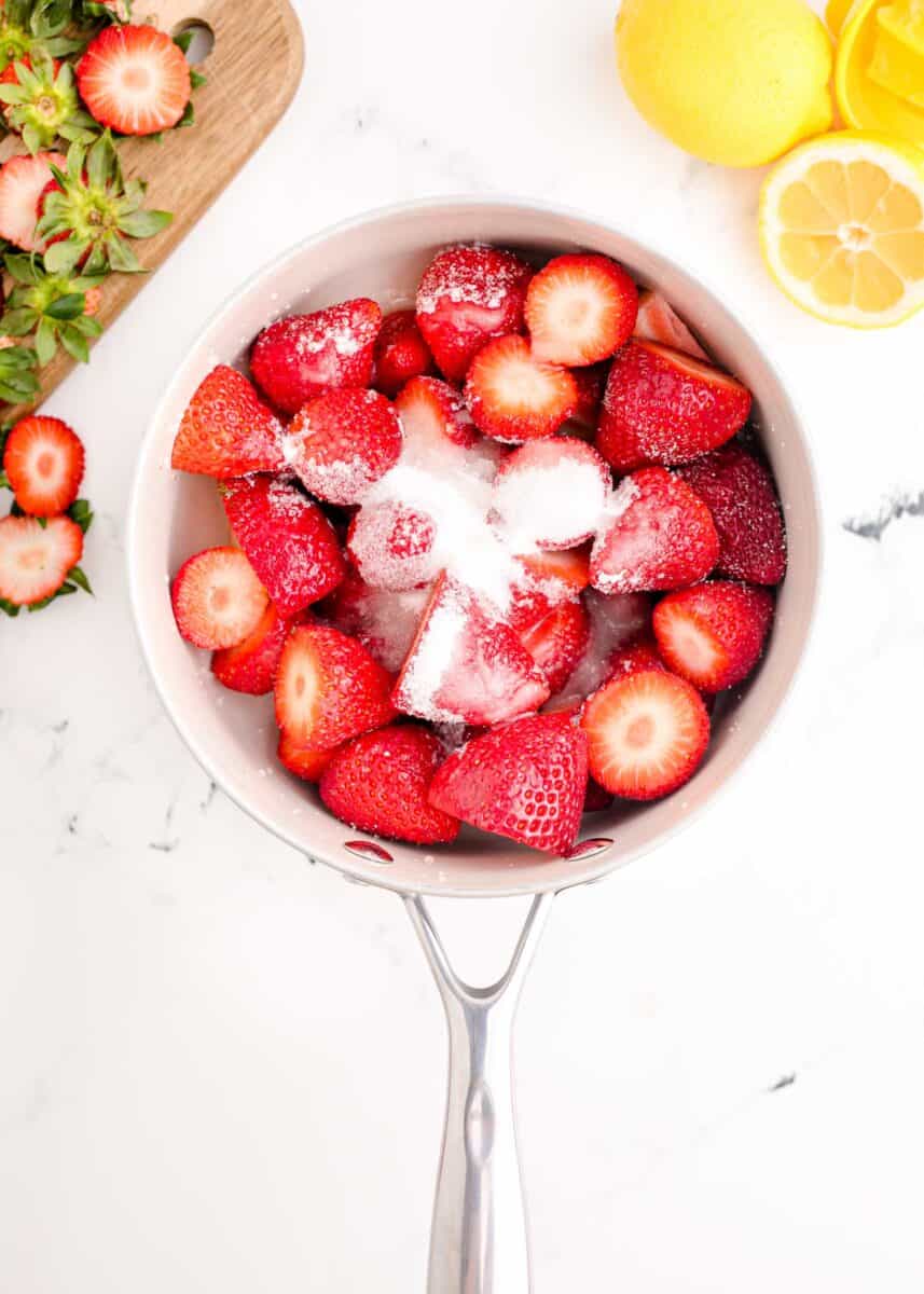 strawberries covered in sugar and lemon juice in a small sauce pan on a marble countertop