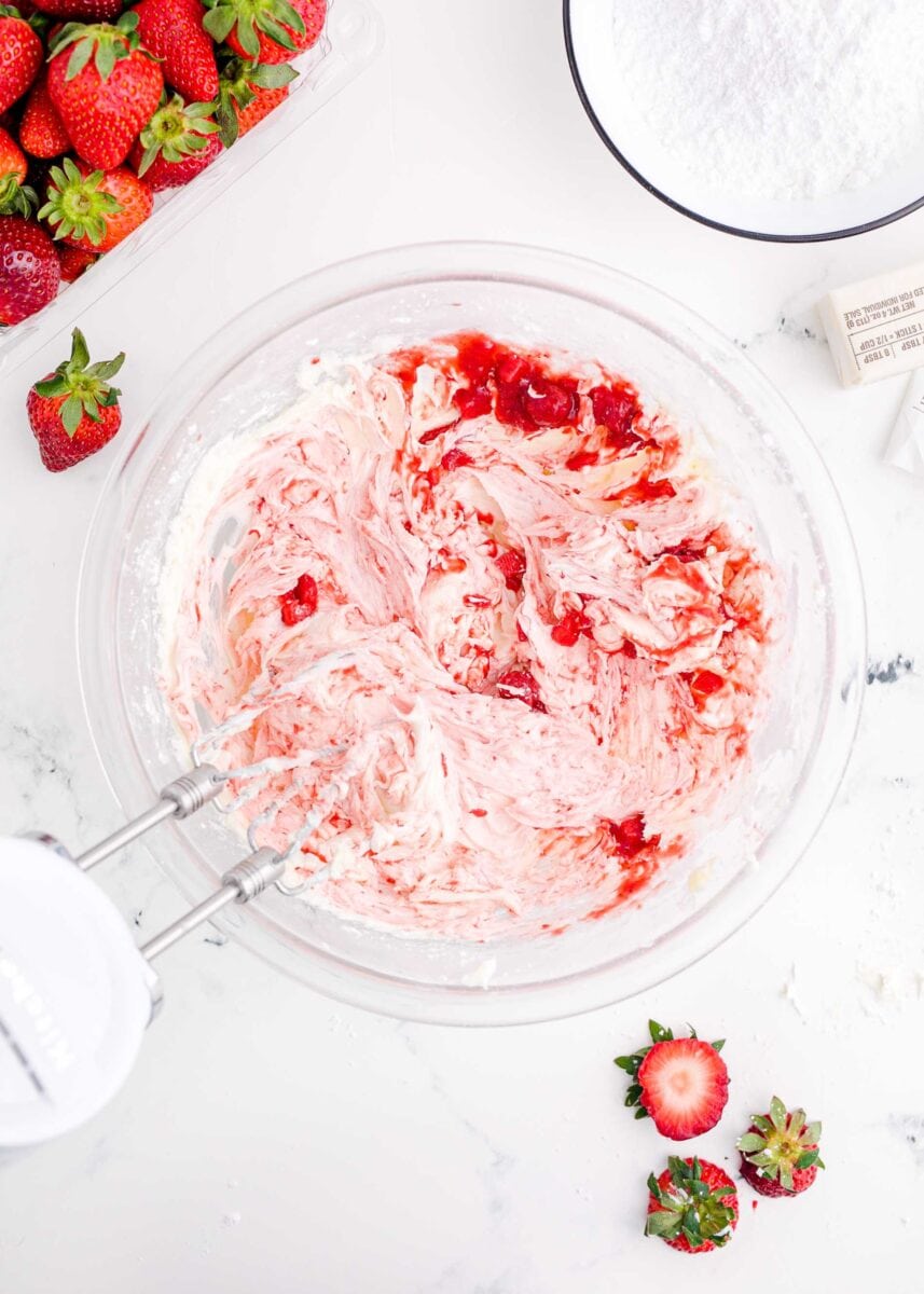 strawberry reduction added to the butter mixture in a clear bowl with a hand mixer in the bowl