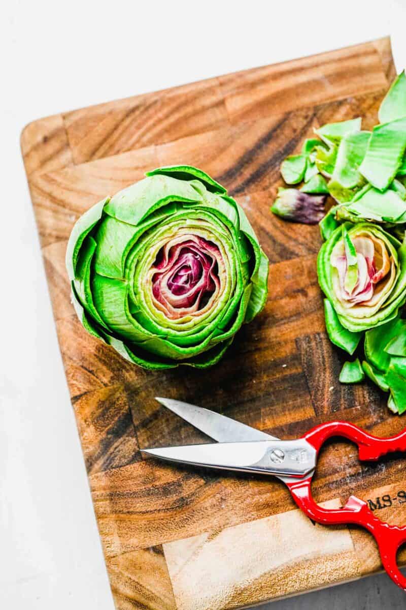 thorny tips of artichoke bud on a wooden cutting board with the tips trimmed off next to a pair of red scissors