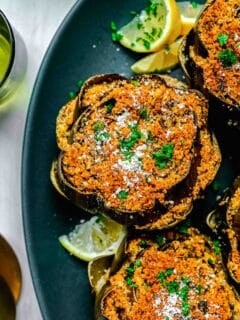 stuffed artichokes on top of a teal platter with fresh lemons throughout and fresh parsley and parmesan cheese sprinkled on top