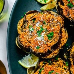 stuffed artichokes on top of a teal platter with fresh lemons throughout and fresh parsley and parmesan cheese sprinkled on top