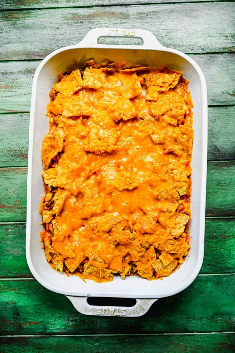 melted cheese on top of crushed up tortilla chips in a white casserole baking dish on a green background