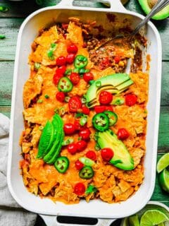 taco casserole in a white casserole baking dish where a spoon shows inside contents. fresh tomatoes, avocado slices, and jalapeno slices are on top