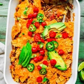 taco casserole in a white casserole baking dish where a spoon shows inside contents. fresh tomatoes, avocado slices, and jalapeno slices are on top