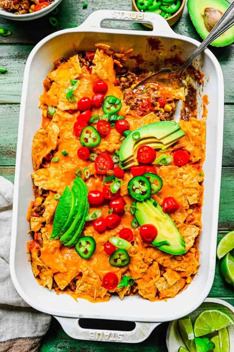 taco casserole in a white casserole baking dish where a spoon shows inside contents. fresh tomatoes, avocado slices, and jalapeno slices are on top