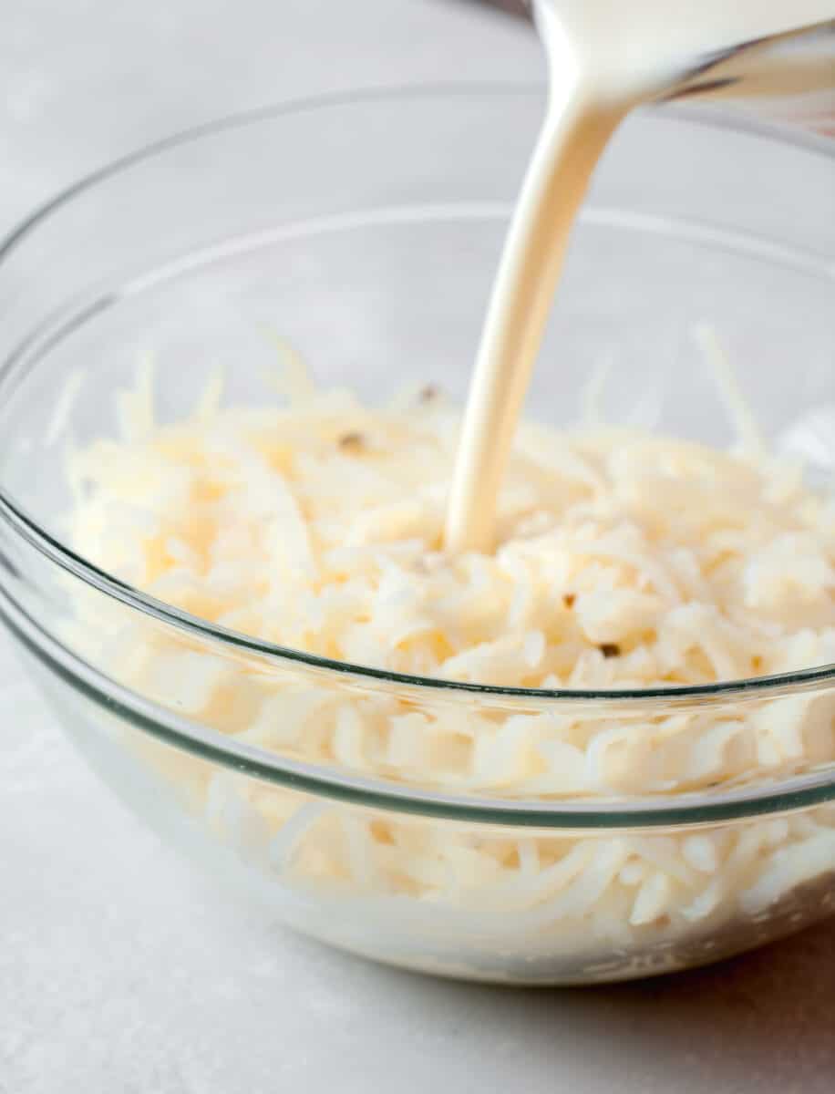 milk and egg mixture being poured into a clear bowl with shredded potatoes