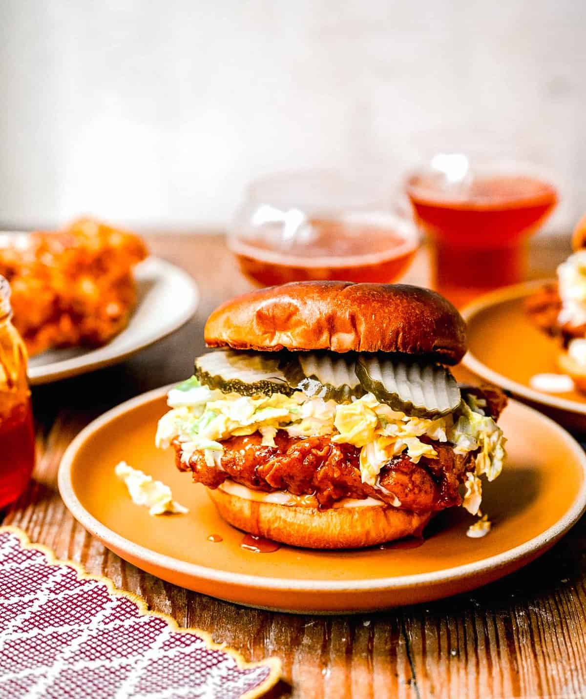 Hot honey chicken sandwiches on plates near a jar of hot honey and 2 glasses full of an beverage.