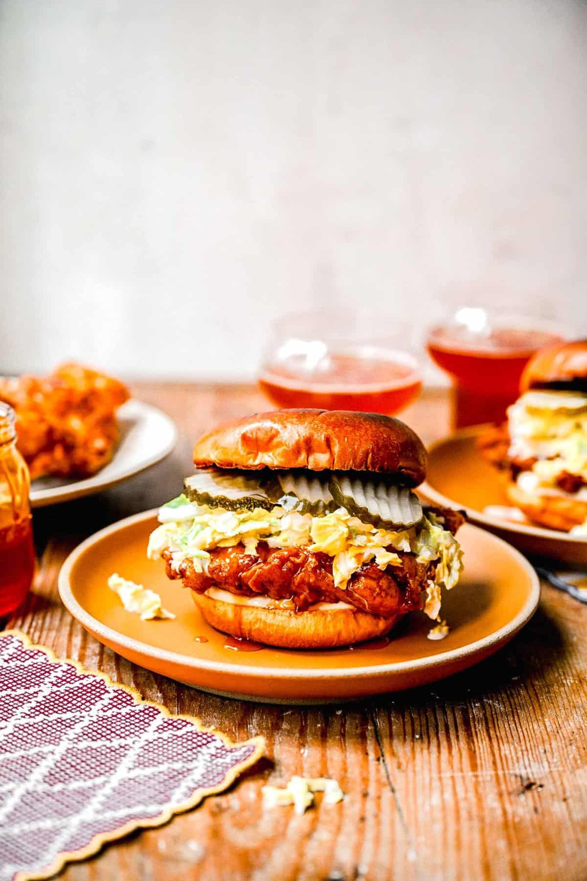Hot honey chicken sandwiches on plates near a jar of hot honey and 2 glasses full of an beverage.
