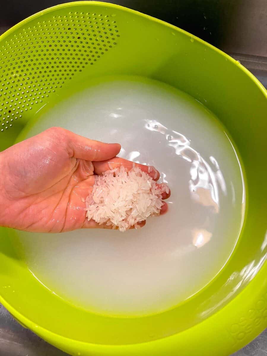 a hand holding washed jasmine rice in a bright green colander with starchy water