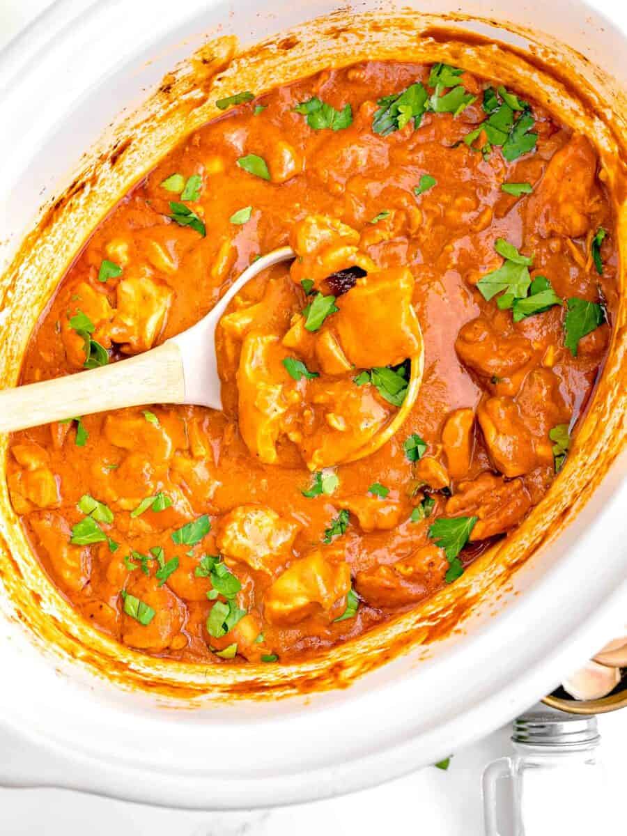 a white ladle is scooping up some butter chicken from the slow cooker.